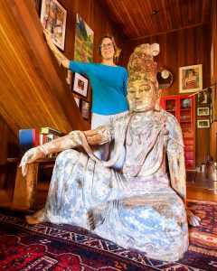 Woman standing behind an old wooden statue of a seated bodhisattva in her home.