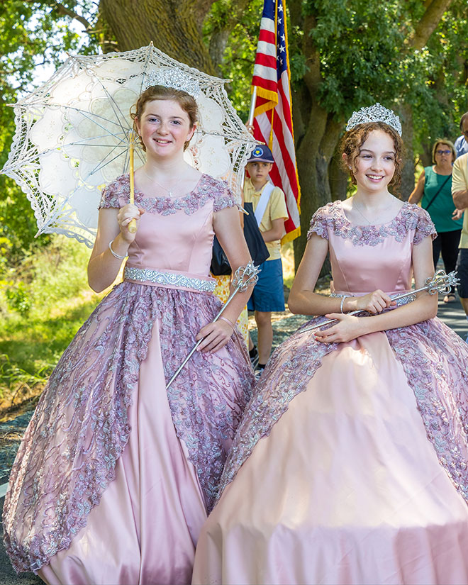 Two young women dressed in ballgowns and tiaras walk in a Festa parade, one carrying a parasol
