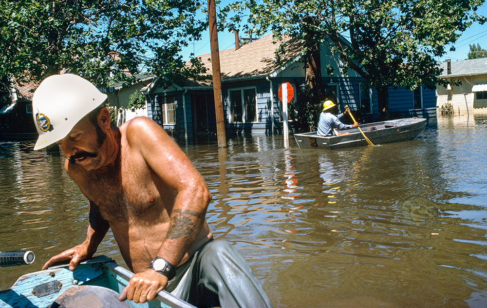 A shirtless man hoists himself into a boat from floodwaters