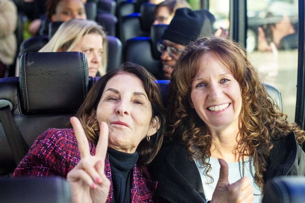 Two women on a bus, one holding up two fingers in a peace or victory sign, one holding her thumb up