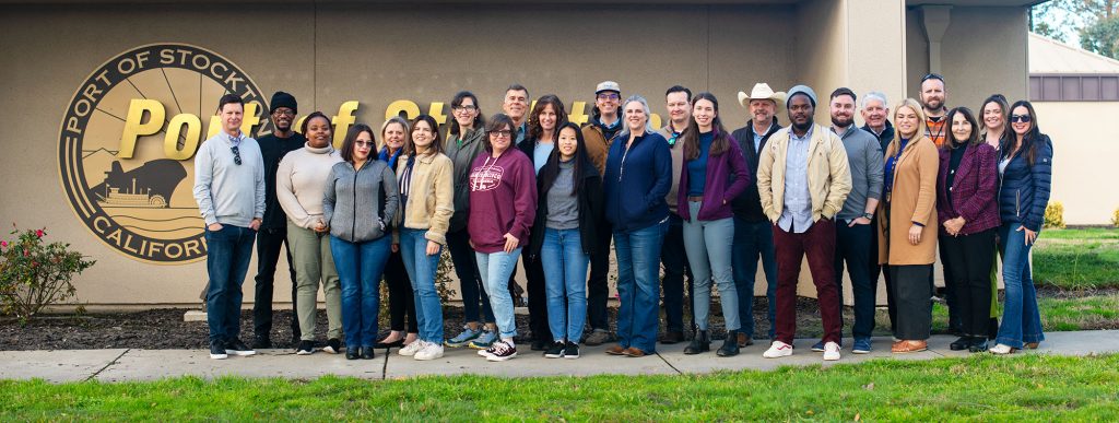 A group photo of adult men and women standing on front of a building with a large Port of Stockton logo on it