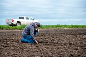 A man holding a coffee cup kneels in tilled soil
