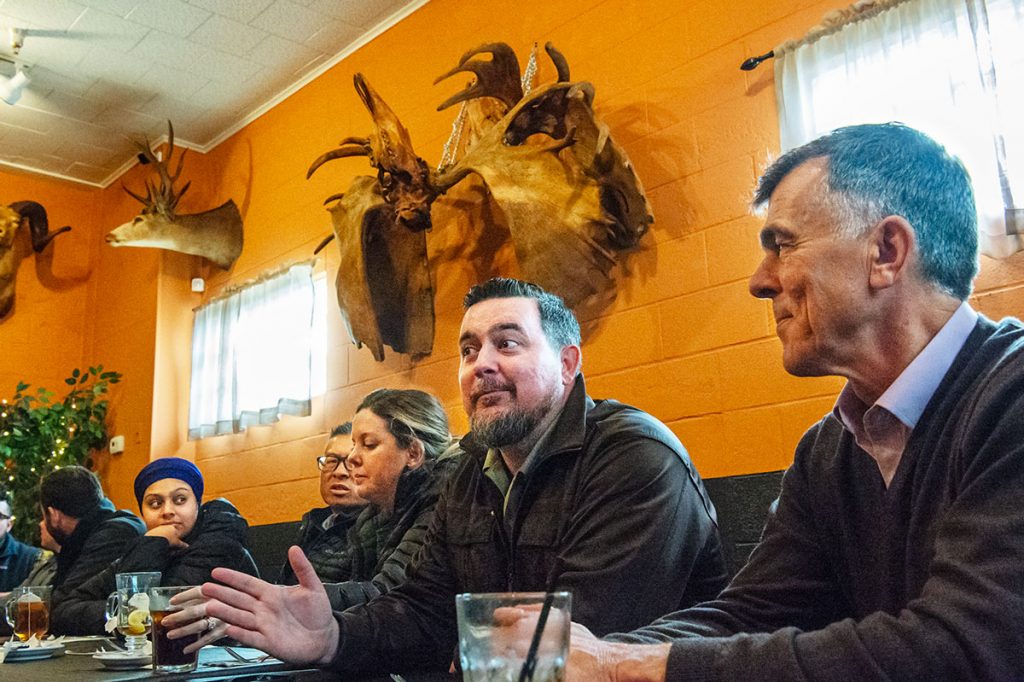 A man at a restaurant table gestures in a conversation with an unseen person across the table; in the background on the wall are the skulls and antlers of two moose that had locked horns.