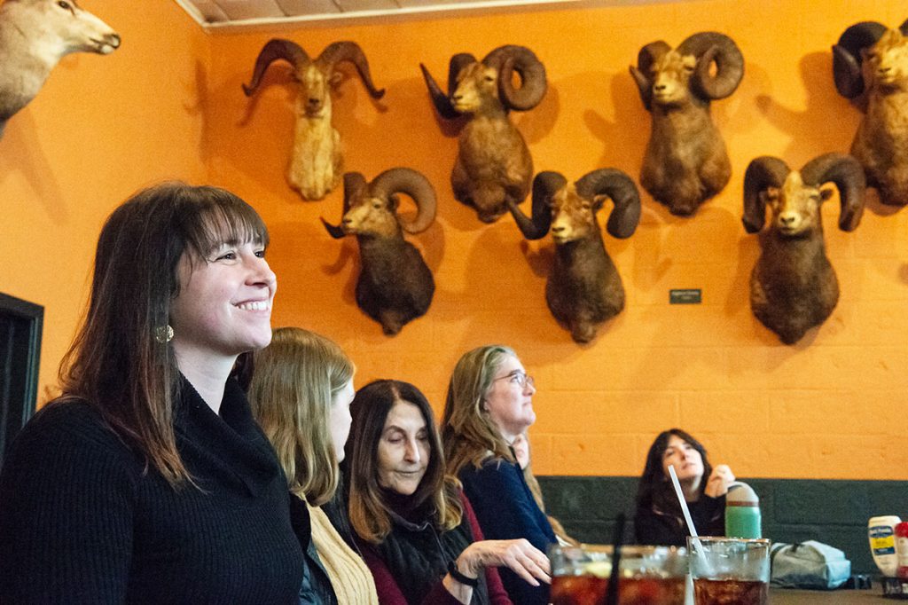 A woman at a restaurant table full of people smiles; on the wall behind them is a wall of taxidermied bighorn sheep heads.