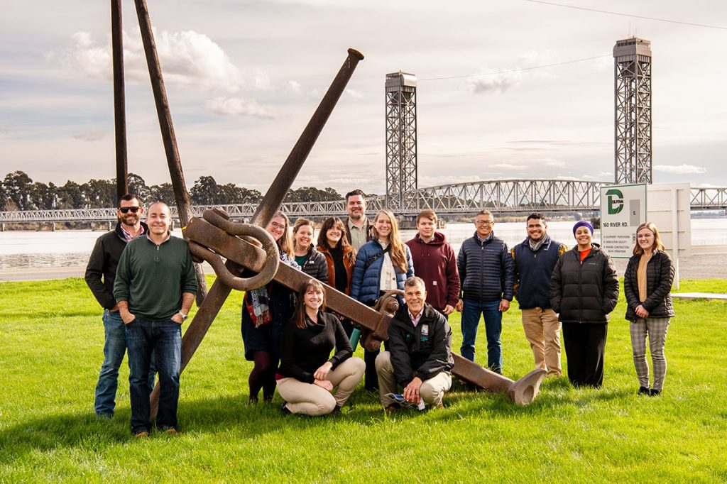 A group poses with a giant anchor on a lawn with a wide river and vertical lift bridge in the background
