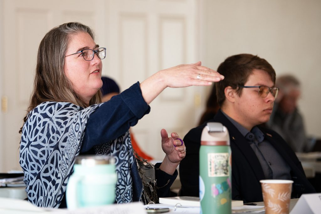 A woman at a conference table gestures in front of her with palm facing down