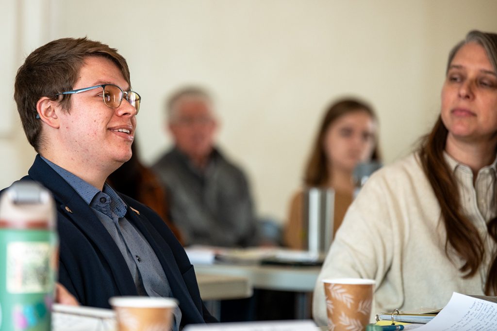 A man at a conference table smiles a a woman seated next to him looks on