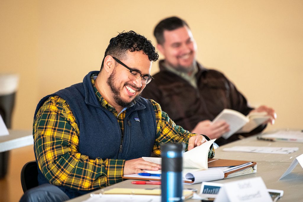 A man at a conference table smiles as a man seated next to him laughs