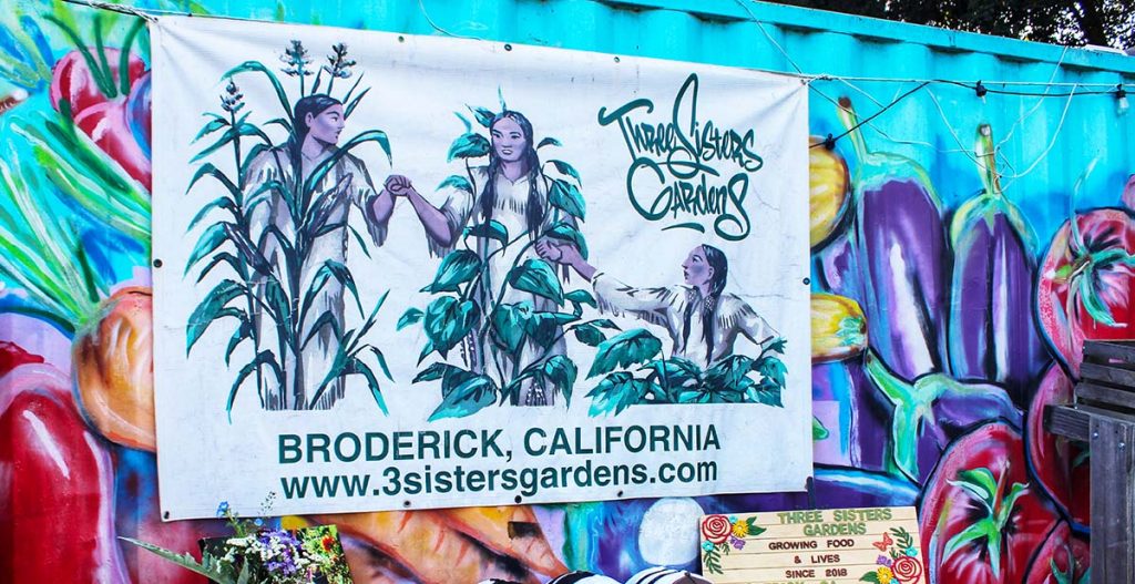 A sign for a community garden, tied to a container painted with a vivid mural of fruits and vegetables.