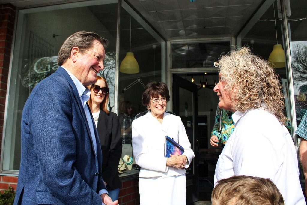Three women chat with a smiling man outside a storefront on a sunny day